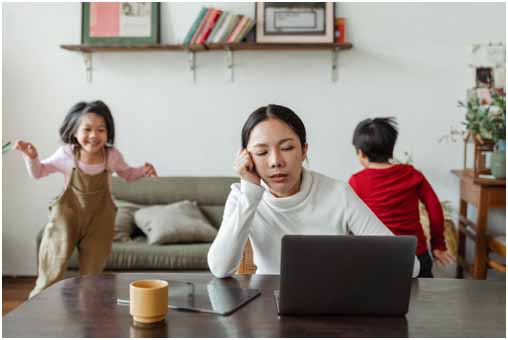 A worn-out mom sitting in front of a laptop with children running in the background.